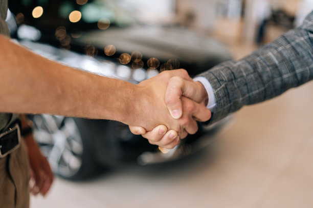 以汽车为背景，在汽车经销店里，穿着西装、面面相认的汽车推销员和快乐的男顾客握手的特写镜头。图片下载