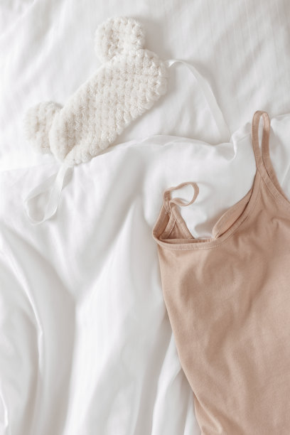 白色床单背景上的米色女式睡衣背心和睡眠眼罩。俯视图舒适舒适的夏天的衣服睡觉躺在枕头和毯子在床上。情调浪漫，生活格调唯美图片下载