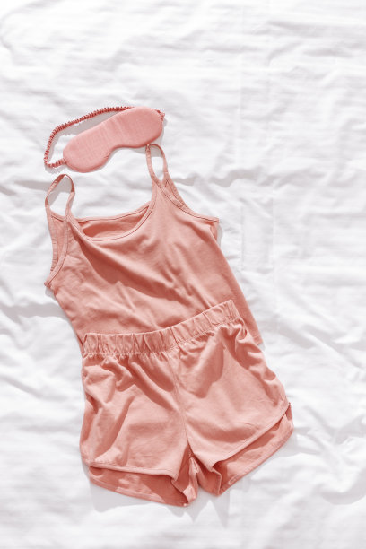 粉色睡衣和眼罩罩在白色皱巴巴的被褥上。舒适的睡衣，晚上舒适休息。单衣，短裤，淡粉色睡眠面膜，睡眠好概念图片下载