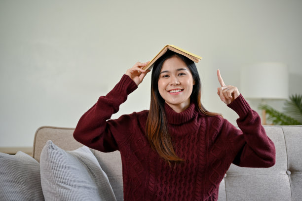 可爱的亚洲女人穿着红色毛衣，坐在沙发上把书放在头上。图片下载
