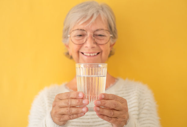 模糊的肖像美丽的白人年老妇女微笑看着满是水在她的手，站在黄色的背景。快乐的老人享受健康的生活方式图片下载