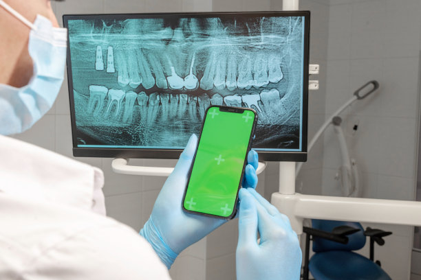 牙科医生手持无菌手套，手持绿色屏幕手机，在牙科诊所的牙齿x光显示器上。正畸医生滚动智能手机色度键屏幕模型模板显示牙齿。图片下载