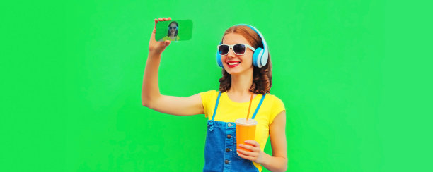 幸福微笑的年轻女子的肖像自拍与智能手机在耳机听音乐的绿色背景图片下载