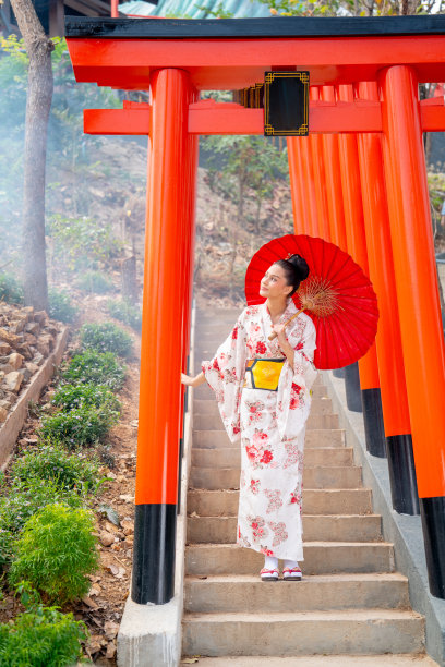穿着日式服装的漂亮女人的垂直形象，拿着红色的雨伞，站在红色拱形入口的楼梯上，幸福地看着左边。图片下载