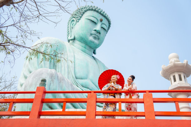 两个亚洲美女的广角镜头，她们站在绿色大佛像底座区域的红色栏杆旁，幸福地望向左边。图片下载