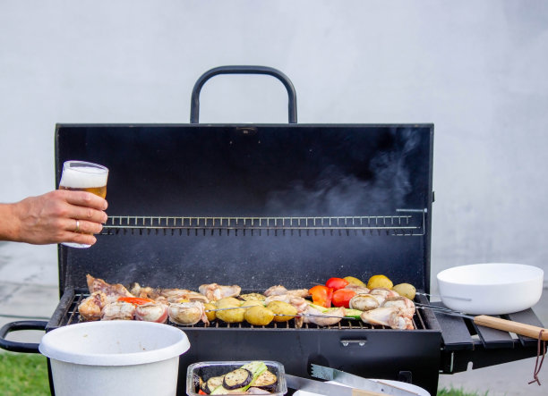 男人手里拿着啤酒，在烤架的背景上放着蔬菜和肉。有选择性的重点。图片下载