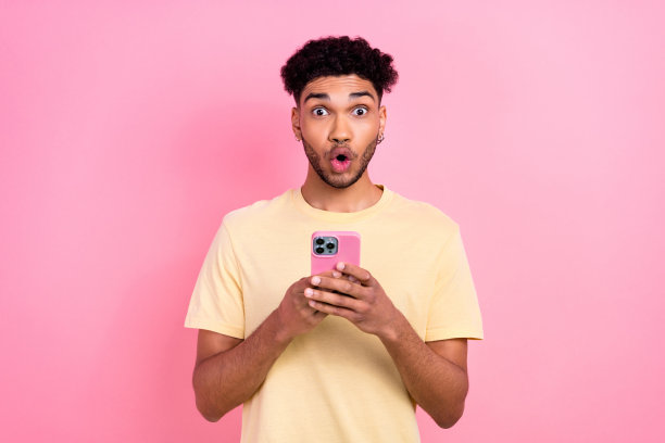 照片中，一个惊讶得说不出话的男孩，留着非洲式发型，穿着黄色t恤，拿着智能手机，撅起嘴唇，孤立在粉红色的背景上图片下载