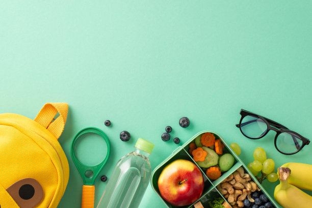 在学校里拥抱健康的生活方式——午餐盒的俯视图，里面有天然的蔬菜，水果，坚果，浆果，水瓶，放大镜，可爱的背包，蓝绿色背景上的眼镜，空白的地方放文字或广告图片下载