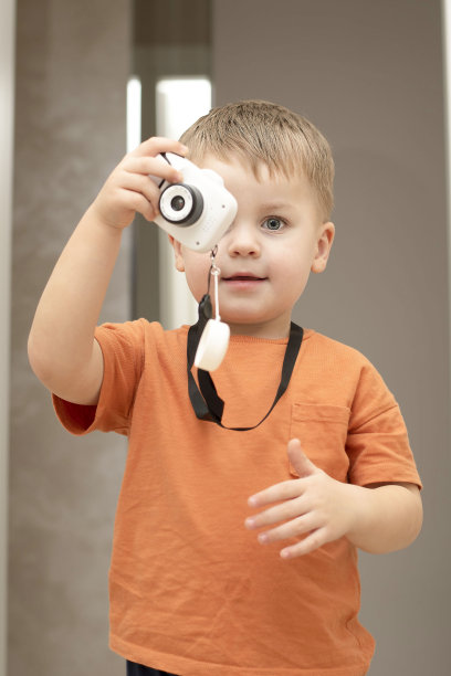 人。帅气、风趣、情绪化的白人金发小男孩穿着橙色t恤，拿着儿童白色相机在家里拍照。图片下载