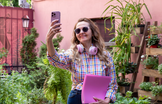 户外照片，快乐的积极时尚的女性，脖子上围着丝巾，在惊人的背景下摆姿势，坐在长凳上，周围是绿色的灌木丛和紫罗兰花，自拍图片下载