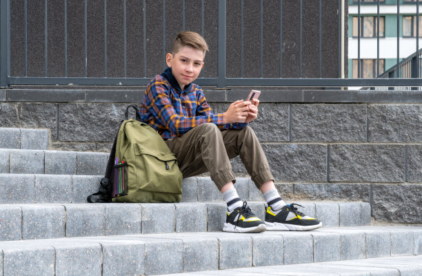 兴高采烈的男学生提着公文包沿街跑去。可爱快乐的小男孩正在用手机看书。回到学校的概念。图片下载