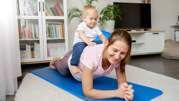 当妈妈在健身训练课上做平板支撑时，可爱的小男孩坐在妈妈的背上。居家保健、运动和瑜伽的概念。图片下载