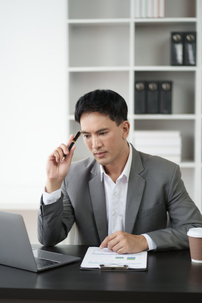 穿着灰色西装的聪明的年轻亚洲商人在办公室里用笔记本电脑工作。图片下载