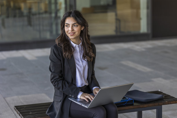 微笑的女商人在笔记本电脑上工作。保险代理人在电脑上查看电子邮件，坐在现代化办公中心附近的户外。图片下载