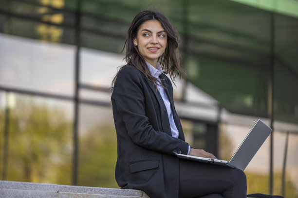 微笑的女商人在笔记本电脑上工作。保险代理人在电脑上查看电子邮件，坐在现代化办公中心附近的户外。图片下载