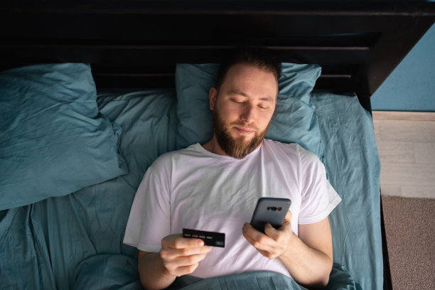 在家网购，安全无忧，远程支付。千禧一代男人拿着智能手机和信用卡，在床上购物图片下载