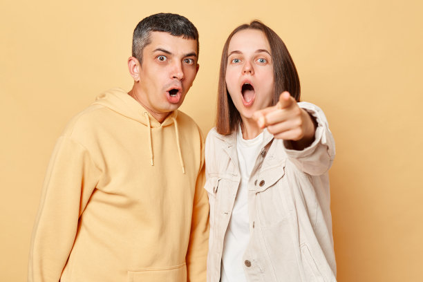 震惊的女人和男人穿着休闲风格的衣服孤立地站在米色的背景下，女人指着镜头，人们用惊讶的大眼睛看着。图片下载