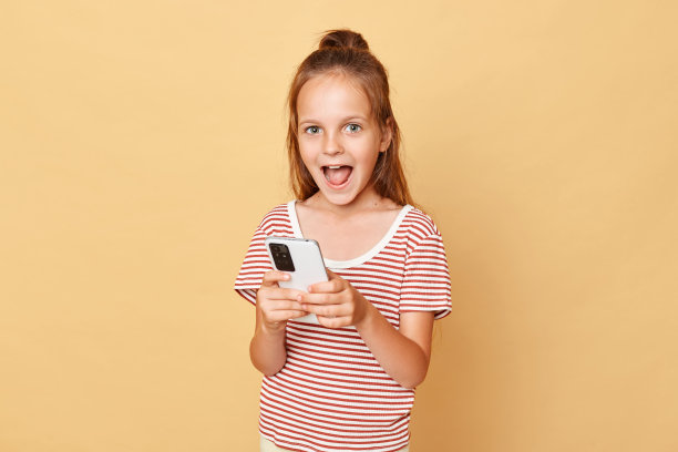 惊讶地笑着，穿着条纹t恤的棕色头发的小女孩站在米色的背景下，拿着手机玩电子游戏，上网冲浪，欢欣鼓舞图片下载