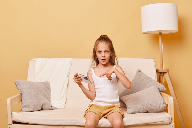 震惊的棕色头发的小女孩用手机指着它的屏幕，用大眼睛和张开的嘴看着相机震惊的内容惊讶的广告。图片下载