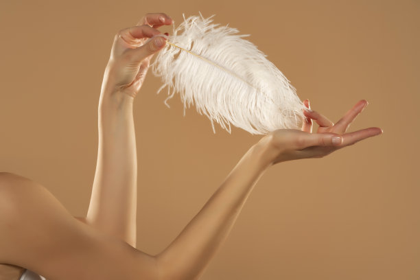 年轻女子的手触摸她的皮肤与白色羽毛在米色背景。美丽身体护理概念图片下载