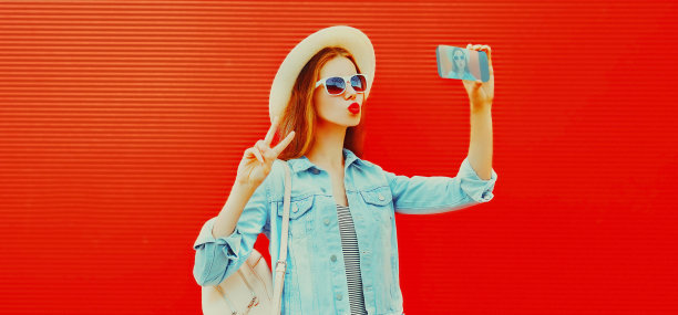 美丽的幸福女人的肖像自拍的智能手机吹她的嘴唇戴夏天草帽，红色背景背包图片下载