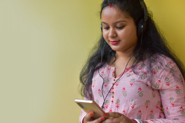 年轻的印度女子一边听音乐，一边用手机，隔绝在黄色背景下。图片下载