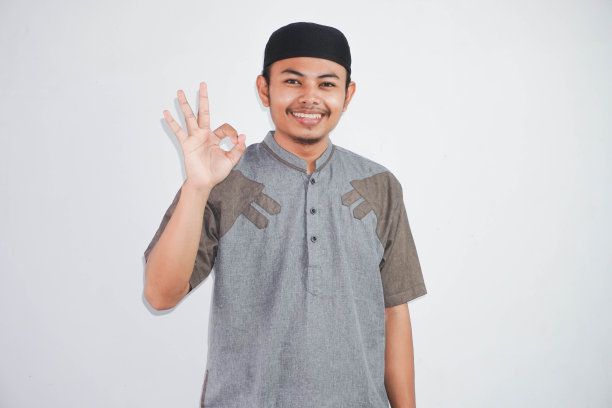 年轻英俊的亚洲穆斯林男子微笑着，用手和手指做着“ok”的手势，穿着灰色的科科衣服，孤立在白色的背景上。成功的表达。图片下载