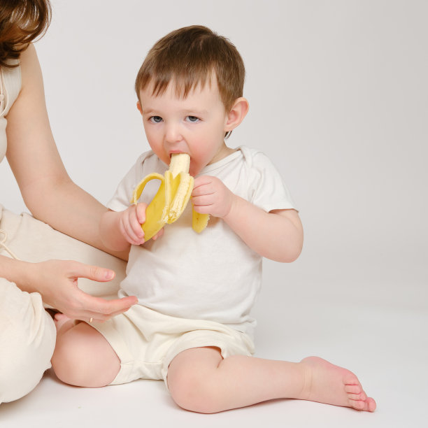 快乐的宝宝和妈妈吃香蕉在工作室白色背景。一个微笑的孩子和妈妈吃水果的照片。2岁左右(1岁9个月)图片下载