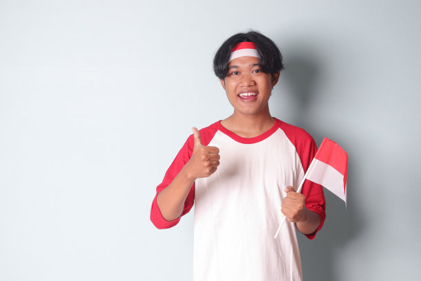 照片中的亚洲男子身穿t恤，头上系着红白丝带，手持印尼国旗，展示产品，手指着远方。灰色背景上的孤立图像图片下载