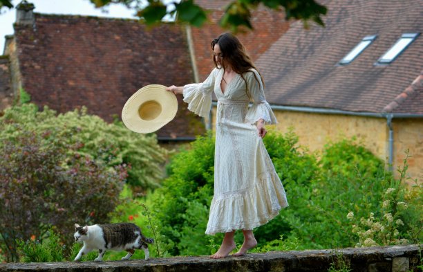 年轻优雅的黑发女子，身穿复古白裙，头戴草帽，在一只猫的陪伴下，面朝绿色自然和典型的多尔多涅河屋顶，走在低矮的墙上图片下载