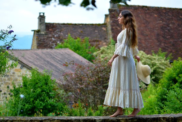 年轻优雅的黑发女子，穿着复古的白色连衣裙，头戴草帽，走在低矮的墙上，面朝绿色的大自然和典型的多尔多涅河的屋顶图片下载