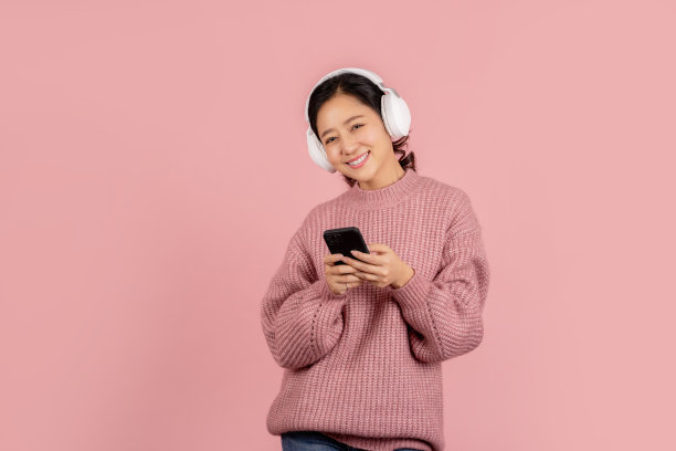 快乐的年轻亚洲女子戴着无线耳机，听着粉色背景的智能手机上的音乐图片下载