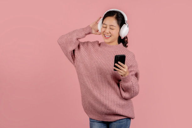 开朗快乐的亚洲女人戴着无线耳机，在粉红色的背景下用智能手机听音乐。图片下载