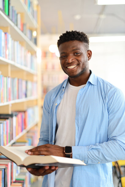一名非洲男子在图书馆里穿着蓝色衬衫，手拿打开的书看手表。背景是摆满各种颜色书籍的书架。图片下载