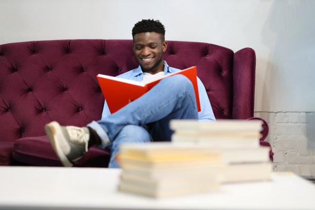 一个黑人坐在紫色沙发上，手里拿着一本红色的书。这个人穿着衬衫和蓝色牛仔裤。图片下载