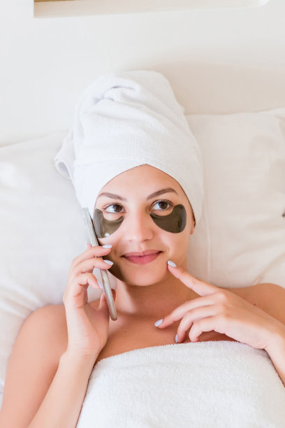 美丽的微笑的年轻女子，戴着眼罩，穿着浴袍躺在床上讲手机。快乐的女孩照顾好自己。美容护肤和健康早晨的概念图片下载