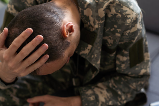 穿着迷彩服的男子坐在白色工作室背景的扶手椅上，用手摸着自己的头，他是一名患有创伤后应激障碍的士兵，服完兵役回到家中图片下载