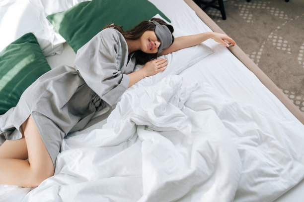 在豪华酒店的卧室里，一个快乐放松的年轻女子戴着睡眠面罩躺在舒适的床上。穿着睡衣的黑发美女肖像图片下载