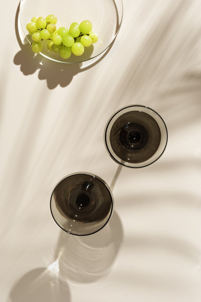 香槟杯着色灰色玻璃和葡萄在米色背景与棕榈叶阴影，从太阳眩光。最小的夏季休息概念。装在彩色玻璃器皿里的汽酒。创造性的俯视图图片下载