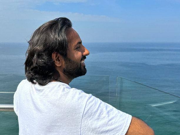 斯里兰卡科伦坡，印度男子站在有玻璃安全屏障的酒店阳台上，靠在扶手上看海景，海景和蓝天背景，侧面视图图片下载