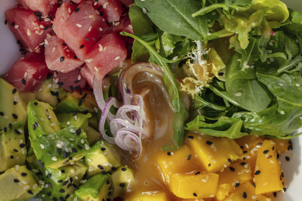 白色桌布上的浅色陶瓷盘子里有新鲜的金枪鱼、牛油果、香草和芒果沙拉。旁边放着一杯白葡萄酒和一碗糖醋酱。图片下载