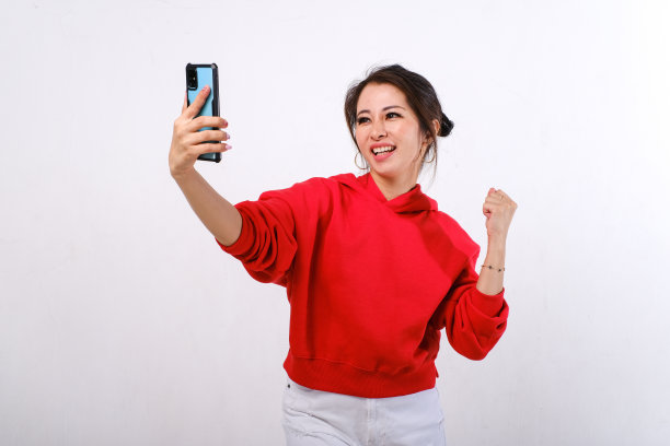 穿着红色毛衣和白色裤子拿着智能手机的兴奋的亚洲女性图片下载