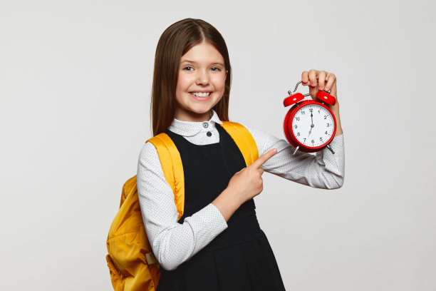 可爱的小女孩穿着校服，背着书包，拿着红色的闹钟指着它图片下载