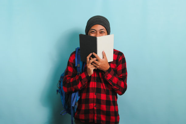 一个年轻的亚洲人站在蓝色的背景下，用一本书遮住脸图片下载