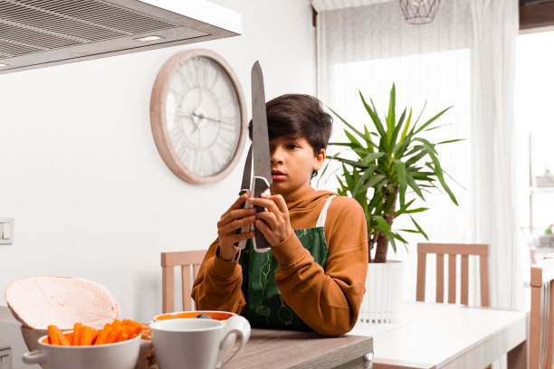 一个10岁的棕色头发的小男孩系着围裙，看着两把刀开始在阳光明媚的厨房里做饭。家庭成员做饭的照片。家庭烹饪图片下载