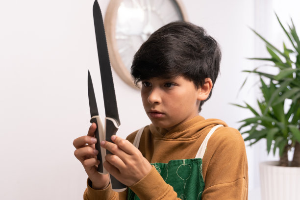 一个10岁的棕色头发的小男孩系着围裙，看着两把刀开始做饭。家庭成员做饭的照片。家庭烹饪图片下载