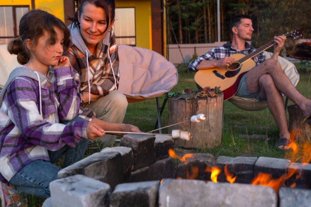 一个穿着格子衬衫的女孩在房子院子里的火上烤棉花糖，父亲在弹吉他。晚上，家人在篝火旁聚会，在旅游椅上野餐图片下载