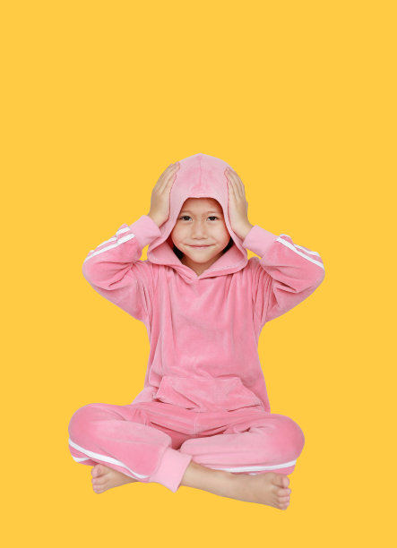 快乐的亚洲小女孩穿着粉红色的运动服或运动服，双手触摸着头巾，独自坐在黄色的背景上。图片下载