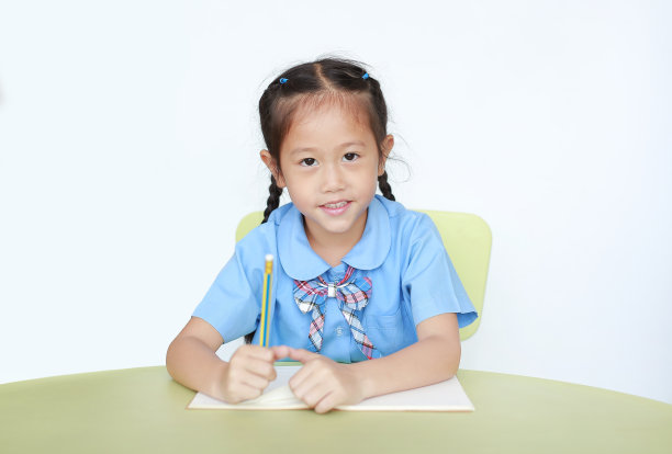 开朗的亚洲小女孩穿着校服，在白底的书桌上写着笔记本。女学生与教育概念。图片下载
