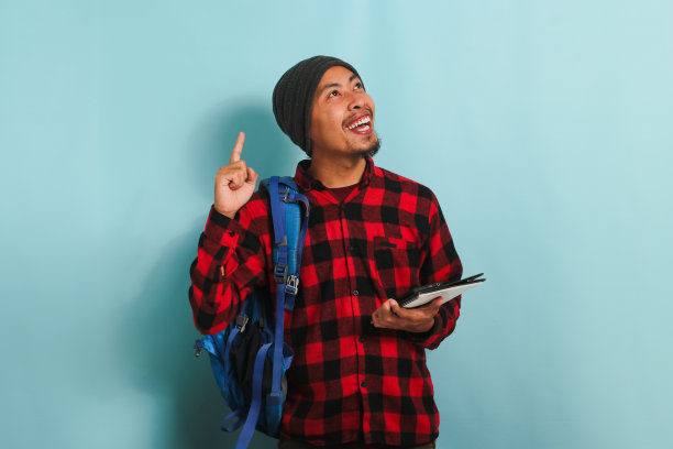 一个兴奋的亚洲年轻人拿着一本书，站在蓝色的背景下，手指着上面图片下载